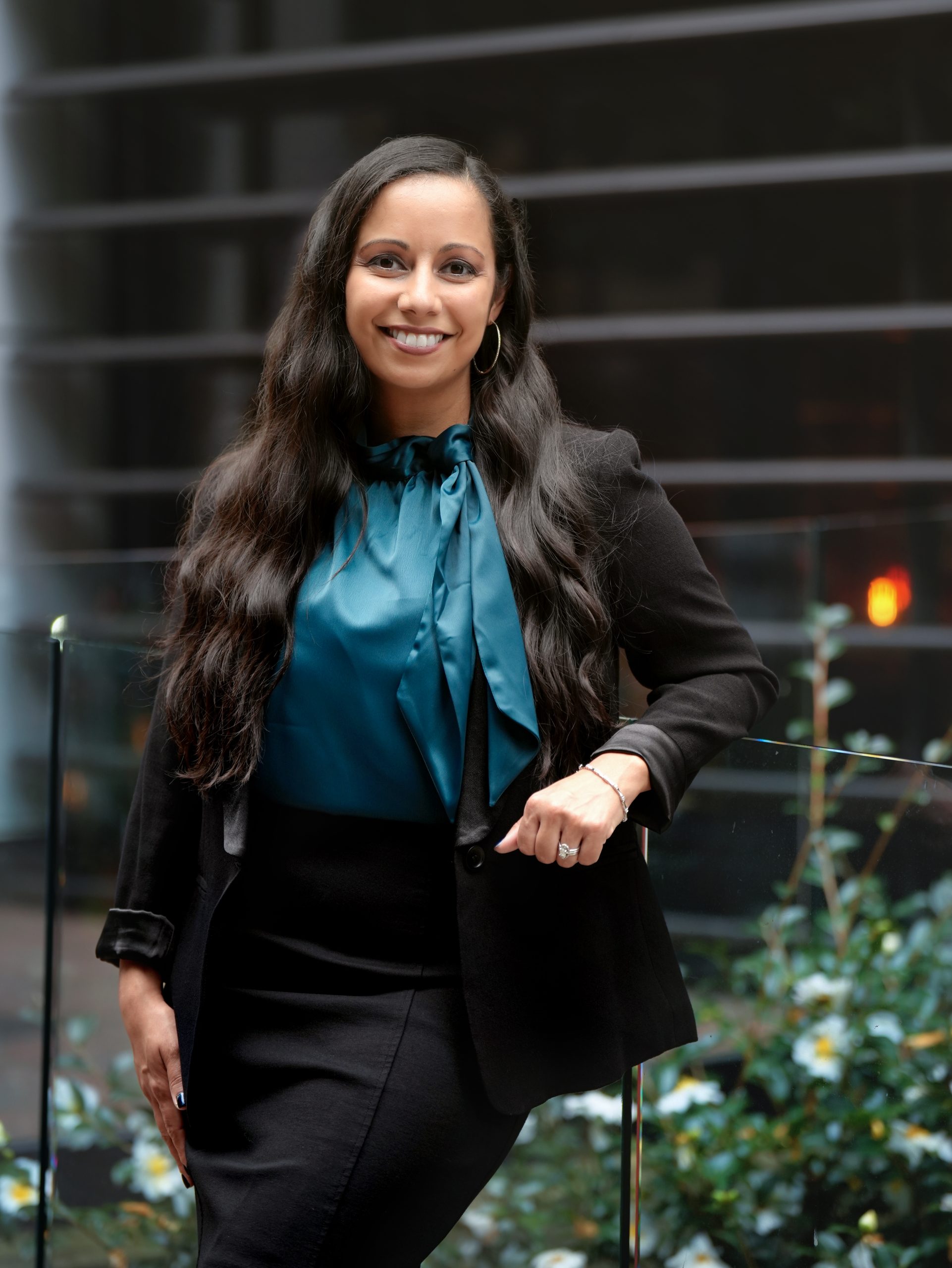 Modern Legal founder and family law attorney Theresa Viera is seen in a teal top and black blazer and skirt smiling for a photo. She is leaning on a clear barrier, with plants and offices from Uptown Charlotte in the background.